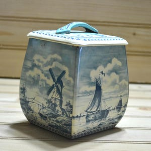 May include: A blue and white tin box with a Dutch windmill and sailboat design. The box has a hinged lid with a blue handle.