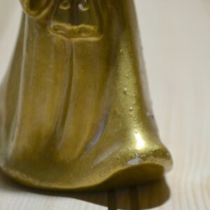 May include: A close-up of a gold-colored figurine with a flowing robe. The figurine has a smooth, reflective surface with some areas showing wear. The base of the figurine is visible, resting on a light-colored wooden surface.