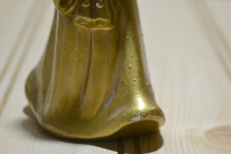 May include: A close-up of a gold-colored figurine with a flowing robe. The figurine has a smooth, reflective surface with some areas showing wear. The base of the figurine is visible, resting on a light-colored wooden surface.