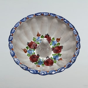 May include: A white ceramic bundt cake pan with a blue and red floral design. The pan has a fluted edge and a central hole in the bottom.
