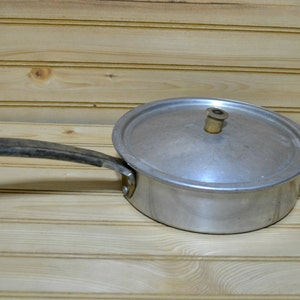 May include: A vintage aluminum saucepan with a lid and a long, dark handle. The lid has a small, light-colored knob. The pan is set against a light wood background.