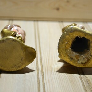 May include: Two vintage figurines, viewed from the bottom, with gold-colored bases and details. One figurine has a visible face with painted features. The other figurine has a large hole in the base.