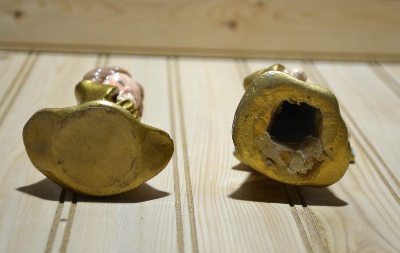 May include: Two vintage figurines, viewed from the bottom, with gold-colored bases and details. One figurine has a visible face with painted features. The other figurine has a large hole in the base.