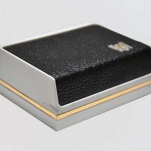 May include: A white and gold rectangular box with a black leather top. The box has a gold trim around the edges.
