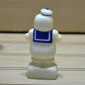 Puede incluir: Una figurita de pl&aacute;stico blanco de un hombre de malvavisco que lleva un sombrero de marinero azul y un cuello de marinero azul y blanco. La figurita est&aacute; de pie sobre una base blanca.