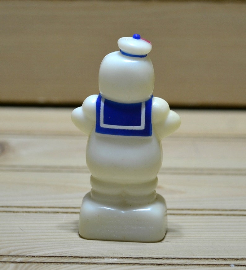 Puede incluir: Una figurita de pl&aacute;stico blanco de un hombre de malvavisco que lleva un sombrero de marinero azul y un cuello de marinero azul y blanco. La figurita est&aacute; de pie sobre una base blanca.