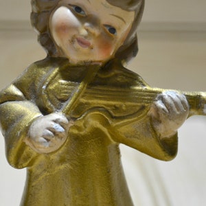 May include: A vintage figurine of a cherubic angel playing a violin. The angel is dressed in a gold-colored robe and has a serene expression. The violin is also gold, with silver accents. The figurine is likely made of ceramic or a similar material.