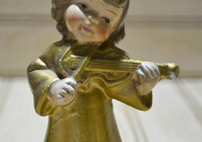 May include: A vintage figurine of a cherubic angel playing a violin. The angel is dressed in a gold-colored robe and has a serene expression. The violin is also gold, with silver accents. The figurine is likely made of ceramic or a similar material.