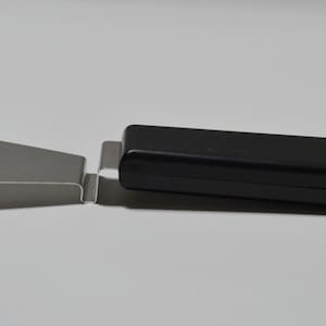 Pode incluir: Uma esp&aacute;tula de bolo de a&ccedil;o inoxid&aacute;vel com cabo preto. A esp&aacute;tula tem uma l&acirc;mina plana e triangular e um cabo longo e retangular. O cabo &eacute; feito de um material liso e brilhante. A esp&aacute;tula &eacute; projetada para servir bolos e tortas.