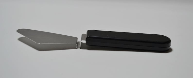 Pode incluir: Uma esp&aacute;tula de bolo de a&ccedil;o inoxid&aacute;vel com cabo preto. A esp&aacute;tula tem uma l&acirc;mina plana e triangular e um cabo longo e retangular. O cabo &eacute; feito de um material liso e brilhante. A esp&aacute;tula &eacute; projetada para servir bolos e tortas.