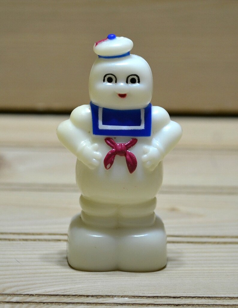 Puede incluir: Una figurita de pl&aacute;stico blanco de un fantasma que lleva un gorro de marinero azul y blanco y una corbata de lazo roja. El fantasma tiene una cara amigable con ojos negros y una boca roja.