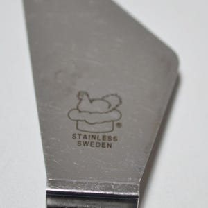 Pode incluir: Servidor de bolo em a&ccedil;o inoxid&aacute;vel com cabo preto. A l&acirc;mina tem uma imagem estampada de uma galinha em uma nuvem e as palavras "STAINLESS SWEDEN".