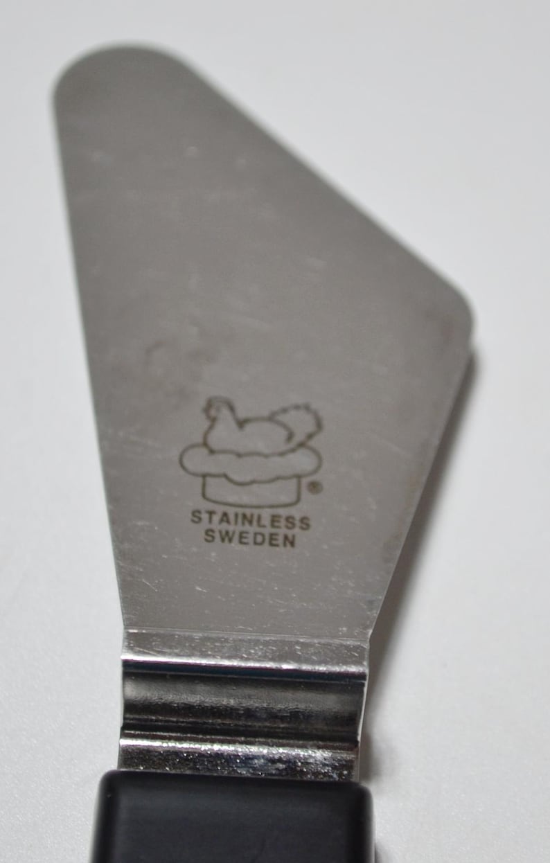 Pode incluir: Servidor de bolo em a&ccedil;o inoxid&aacute;vel com cabo preto. A l&acirc;mina tem uma imagem estampada de uma galinha em uma nuvem e as palavras "STAINLESS SWEDEN".