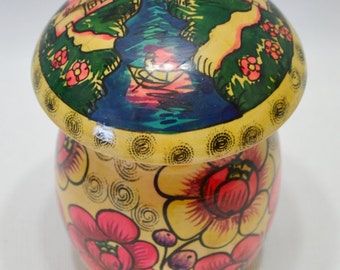 May include: A wooden painted trinket box with a mushroom shape. The lid features a painted scene of a river, trees, houses, and a boat. The body of the box is painted with yellow, pink, and red flowers.