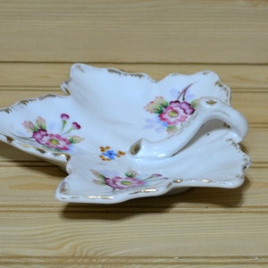 May include: A white porcelain leaf-shaped dish with a handle. The dish is decorated with pink, purple, and blue flowers and gold trim.