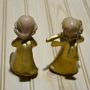 May include: Two small, gold-colored angel figurines with light brown hair. One angel is kneeling, and the other is playing a flute. The figurines are on a light-colored wooden surface. The angels are wearing gold robes.