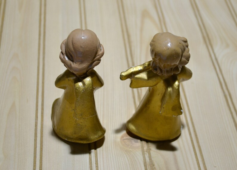 May include: Two small, gold-colored angel figurines with light brown hair. One angel is kneeling, and the other is playing a flute. The figurines are on a light-colored wooden surface. The angels are wearing gold robes.