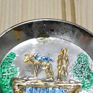Puede incluir: Un cenicero de metal plateado con una imagen colorida de un hombre en un burro con el texto "Knott's" en la parte inferior.