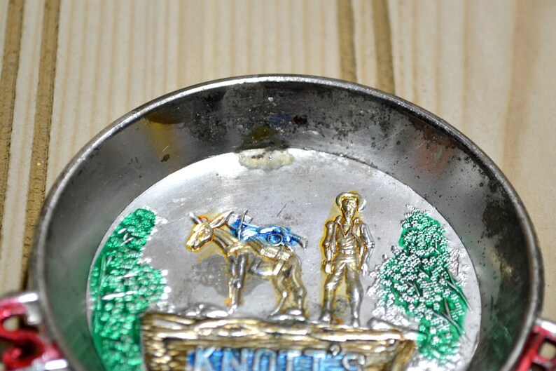 Puede incluir: Un cenicero de metal plateado con una imagen colorida de un hombre en un burro con el texto "Knott's" en la parte inferior.