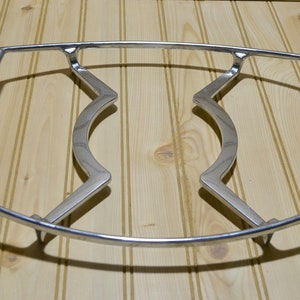 May include: A silver metal hot plate stand with a curved design. The stand has two raised sections in the middle and a wire frame around the edges.
