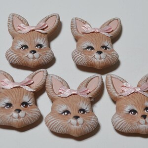 May include: Five ceramic bunny head ornaments. Each bunny has a light brown face with pink ears and a pink bow. The bunnies have black eyes and white eyelashes. The ornaments are suitable for Easter or spring decor.