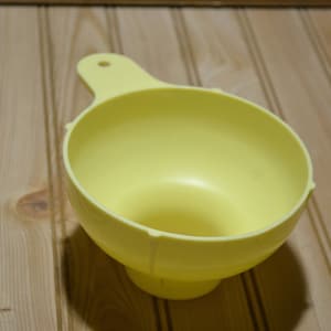 May include: A yellow plastic funnel with a wide top and a narrow spout. The funnel is sitting on a wooden surface.