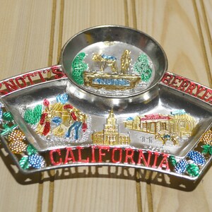 Puede incluir: Una bandeja de recuerdo de metal plateado con un peque&ntilde;o taz&oacute;n en el centro. La bandeja tiene un borde rojo, blanco y azul con un dise&ntilde;o de uvas y hojas. La bandeja tiene las palabras "Knott's Berry Farm California" impresas en ella. El taz&oacute;n tiene una imagen de un camello y un carro con la palabra "Knott's" impresa en &eacute;l.
