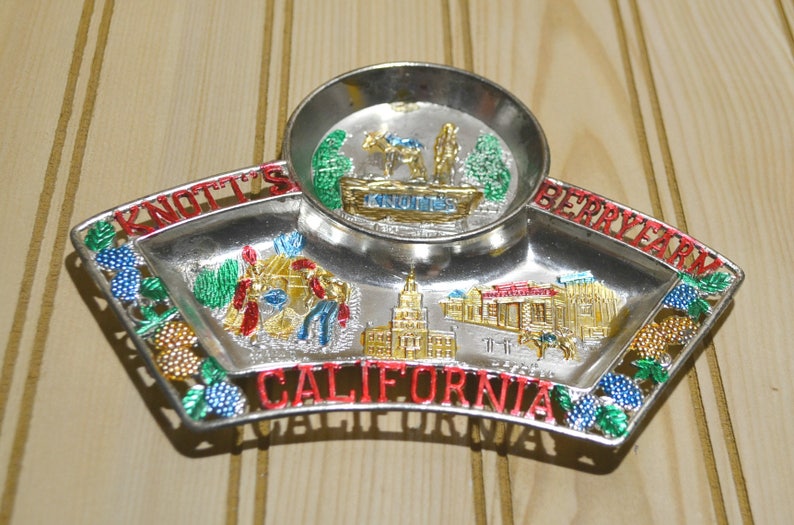 Puede incluir: Una bandeja de recuerdo de metal plateado con un peque&ntilde;o taz&oacute;n en el centro. La bandeja tiene un borde rojo, blanco y azul con un dise&ntilde;o de uvas y hojas. La bandeja tiene las palabras "Knott's Berry Farm California" impresas en ella. El taz&oacute;n tiene una imagen de un camello y un carro con la palabra "Knott's" impresa en &eacute;l.