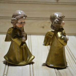 May include: Two vintage angel figurines. The angels have gold-colored robes and wings, with light brown hair and skin. One angel holds a candle. The figurines are on a light-colored wooden surface.