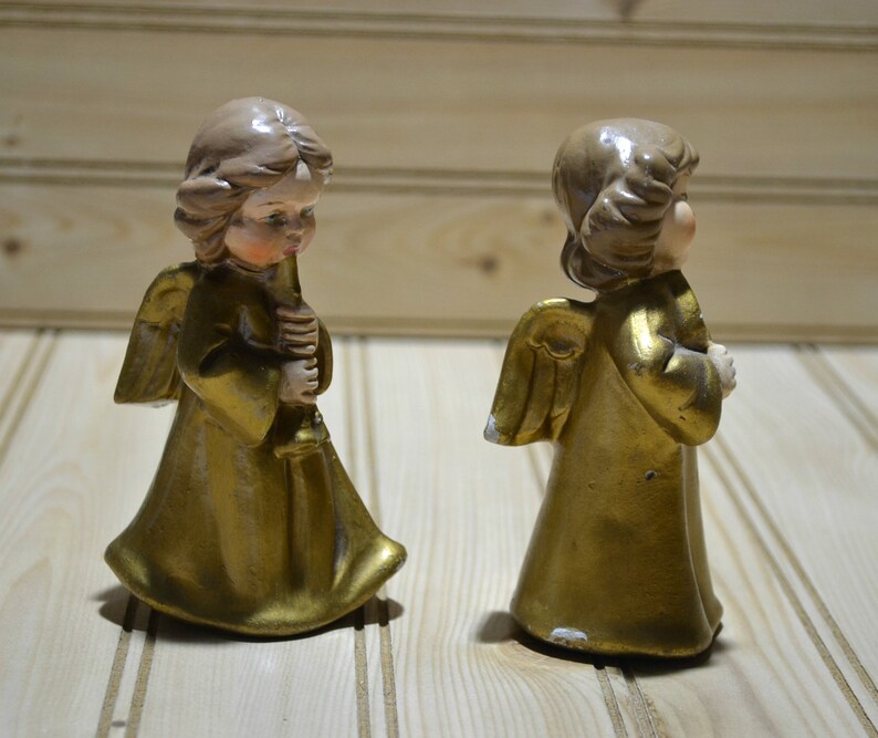 May include: Two vintage angel figurines. The angels have gold-colored robes and wings, with light brown hair and skin. One angel holds a candle. The figurines are on a light-colored wooden surface.