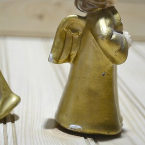 May include: Two vintage gold-painted angel figurines. The angels have detailed wings and are holding objects. The paint shows signs of wear, adding to their antique charm. The figurines are set against a light-colored wooden background.