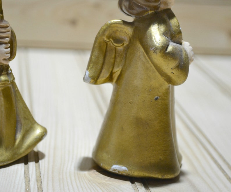 May include: Two vintage gold-painted angel figurines. The angels have detailed wings and are holding objects. The paint shows signs of wear, adding to their antique charm. The figurines are set against a light-colored wooden background.