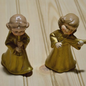 May include: Two ceramic angel figurines. One angel plays a flute, the other a violin. Both angels wear gold robes and have light brown hair. The figurines are on a light wood surface.