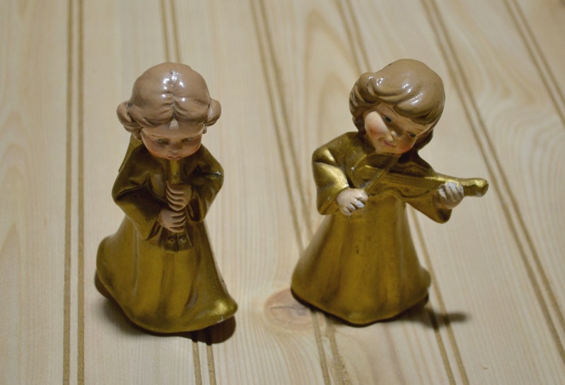 May include: Two ceramic angel figurines. One angel plays a flute, the other a violin. Both angels wear gold robes and have light brown hair. The figurines are on a light wood surface.