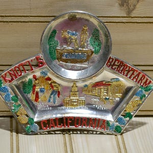 Puede incluir: Una bandeja de recuerdo de metal plateado con un dise&ntilde;o rojo, blanco y azul. La bandeja presenta las palabras "Knott's Berry Farm California" y una representaci&oacute;n de un edificio con un techo rojo.