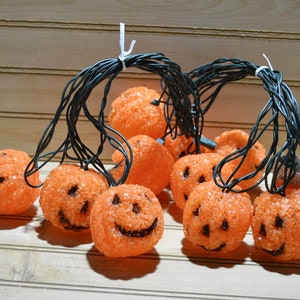 May include: A string of orange pumpkin-shaped lights with black wire. The pumpkins have black painted faces and a textured surface.