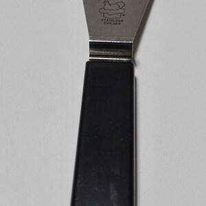 Pode incluir: Um espalhador de a&ccedil;o inoxid&aacute;vel com cabo preto. A l&acirc;mina &eacute; triangular e estampada com "STAINLESS SWEDEN" e um log&oacute;tipo. O cabo tem cerca de 10 cm de comprimento.