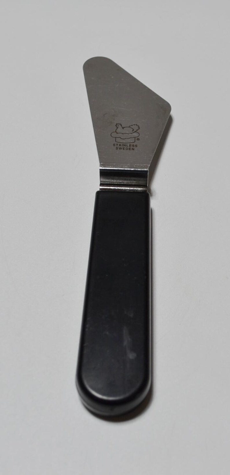 Pode incluir: Um espalhador de a&ccedil;o inoxid&aacute;vel com cabo preto. A l&acirc;mina &eacute; triangular e estampada com "STAINLESS SWEDEN" e um log&oacute;tipo. O cabo tem cerca de 10 cm de comprimento.