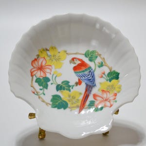 May include: A white porcelain seashell dish with a gold stand. The dish features a colorful parrot with red, blue, and yellow feathers, surrounded by green leaves and yellow and pink flowers.