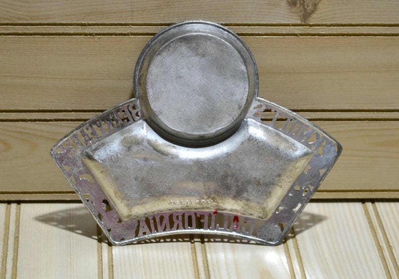 Puede incluir: Un plato de metal plateado con un centro redondo elevado y un borde festoneado. El plato tiene las palabras "California Souvenir" alrededor del borde.