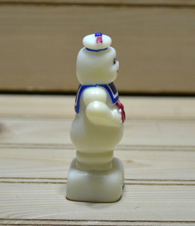 Puede incluir: Una figurita de pl&aacute;stico blanco de un fantasma con un sombrero de marinero azul y rojo y una camisa de marinero azul y blanca. El fantasma est&aacute; de pie sobre una base blanca.