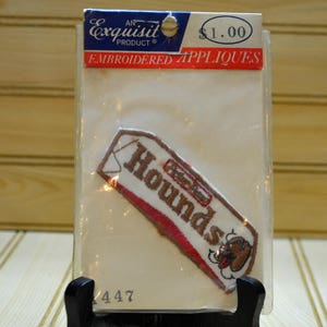 May include: A white package with a brown and red embroidered patch that says "Hound's" with a cartoon dog on it. The package is labeled "An Exquisite Product" and "Embroidered Appliques".