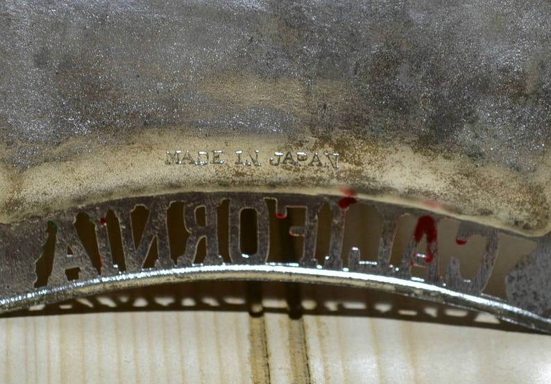 Puede incluir: Una placa de matr&iacute;cula de California de metal plateado con el texto "MADE IN JAPAN" en la parte superior. La placa tiene un dise&ntilde;o recortado de la palabra "CALIFORNIA".