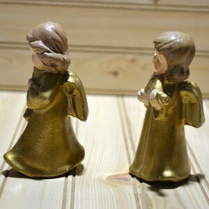 May include: Two gold-colored angel figurines with detailed wings and flowing robes. The angels have light brown hair and are holding a small object. The figurines are standing on a light-colored wooden surface.