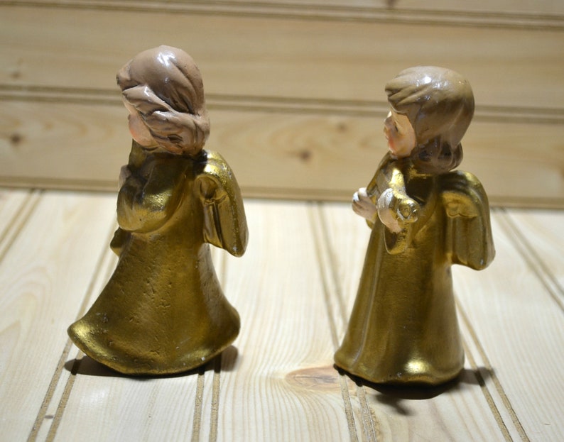 May include: Two gold-colored angel figurines with detailed wings and flowing robes. The angels have light brown hair and are holding a small object. The figurines are standing on a light-colored wooden surface.
