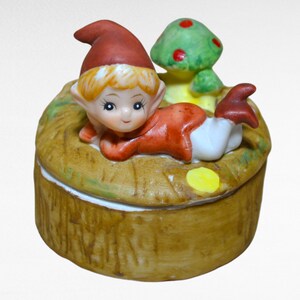 May include: A ceramic trinket box with a whimsical design featuring a red-hatted elf laying on a bed of straw with a green mushroom beside him. The box is decorated with brown, green, yellow, and white paint.