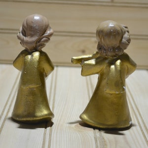 May include: Two small, vintage angel figurines. The angels have gold-colored robes and light brown hair. One angel has its arms outstretched. The figurines are on a light-colored wooden surface.