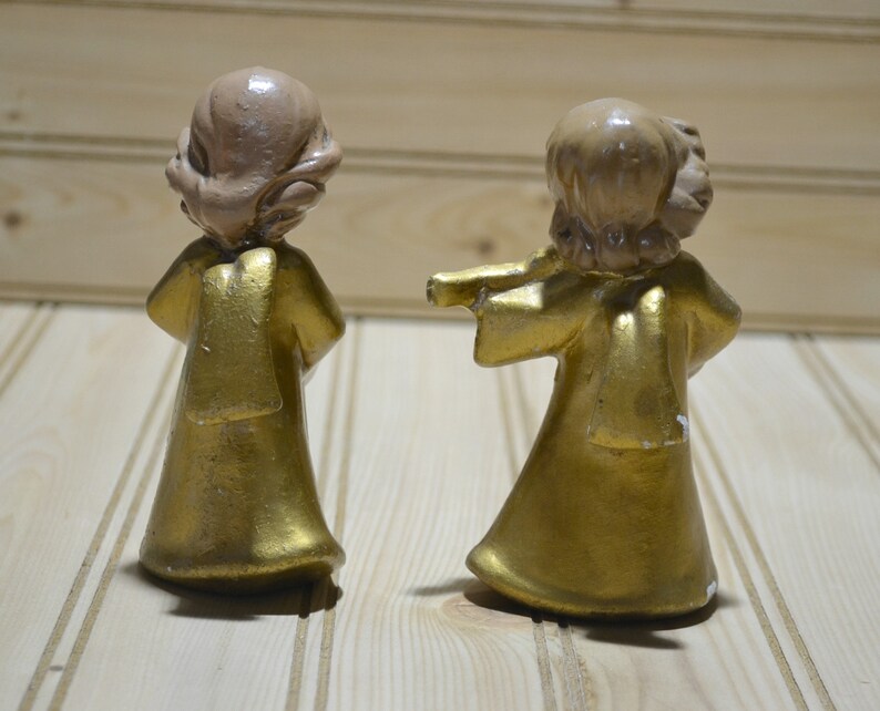 May include: Two small, vintage angel figurines. The angels have gold-colored robes and light brown hair. One angel has its arms outstretched. The figurines are on a light-colored wooden surface.
