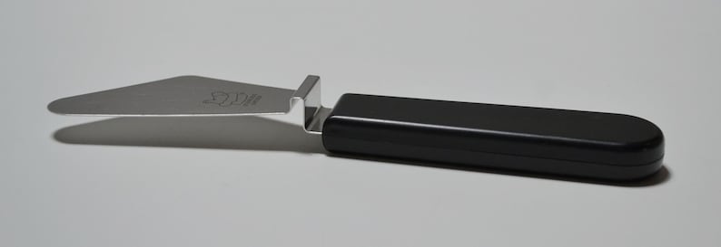 Pode incluir: Uma esp&aacute;tula para bolos com l&acirc;mina de a&ccedil;o inoxid&aacute;vel e cabo preto. A esp&aacute;tula tem uma l&acirc;mina plana e angulada e um cabo preto retangular. A l&acirc;mina tem um pequeno logotipo gravado. Projetada para cortar e servir bolos.