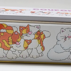 May include: A rectangular white metal box with a lid, featuring cartoon cat illustrations in orange, yellow, and white. The lid has the word "COUPONS" printed on it. The box has a gold-colored trim.